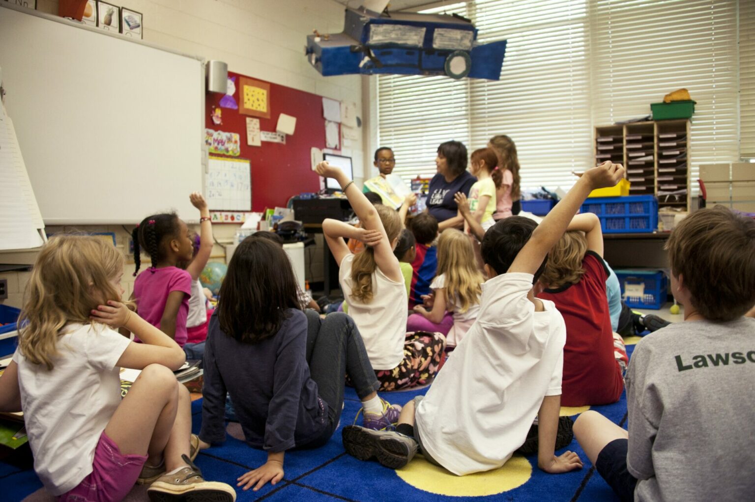 a picture of children sitting on the floor and raising hands for discussion, in a colourful classroom