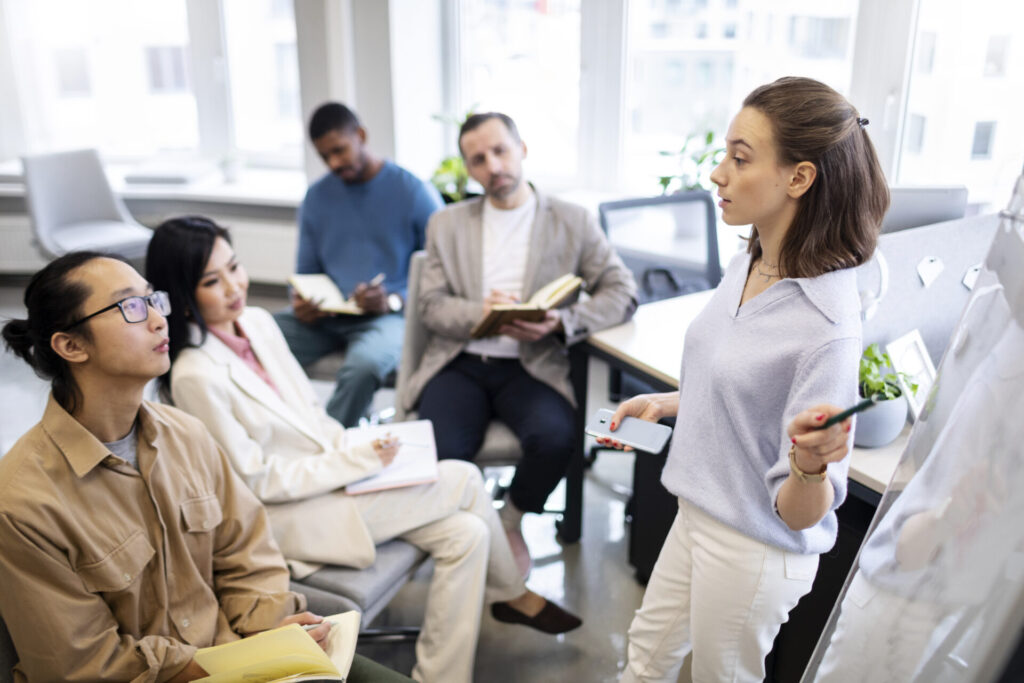 a young woman pointing at a whiteboard while other people in an office setting listen to her