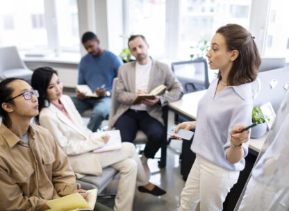 a young woman pointing at a whiteboard while other people in an office setting listen to her